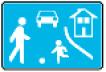 Ein blaues rechteckiges Verkehrszeichen, im Vordergrund sind eine große und eine kleine Person mit einem Ball, im Hintergrund ein Auto und ein Gebäude
